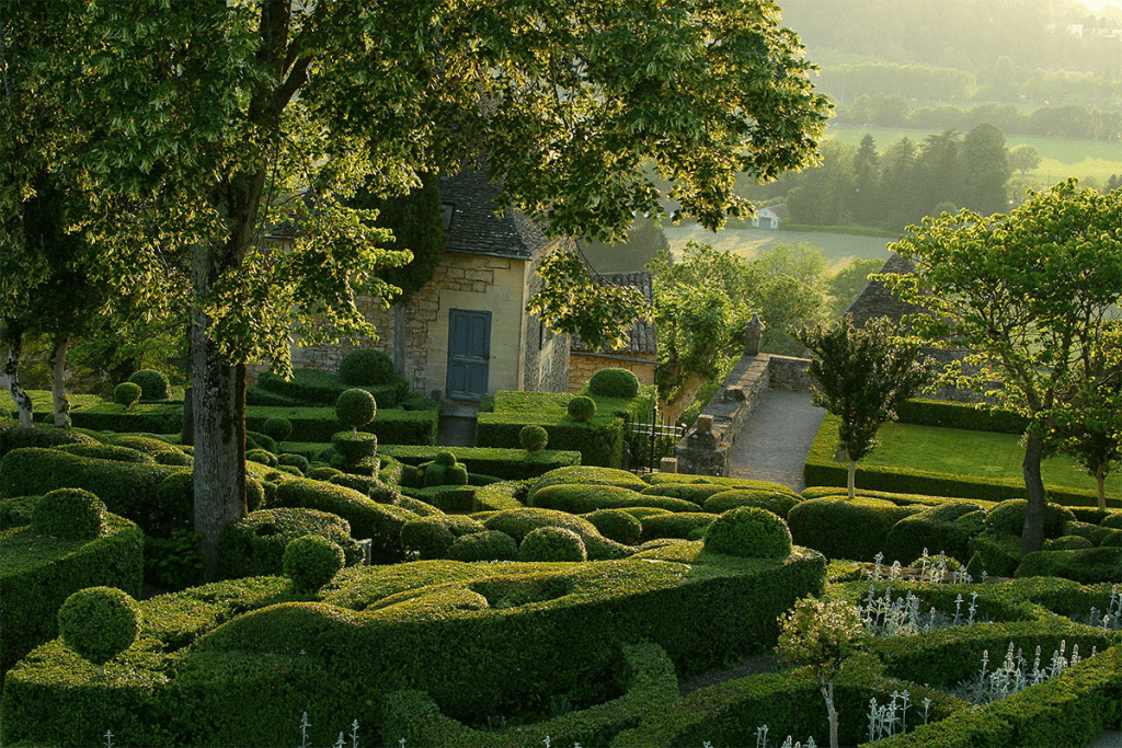 Marqueyssac Gardens in the Dordogne Valley with manicured boxwood hedges, a stone cottage, and lush greenery at sunset