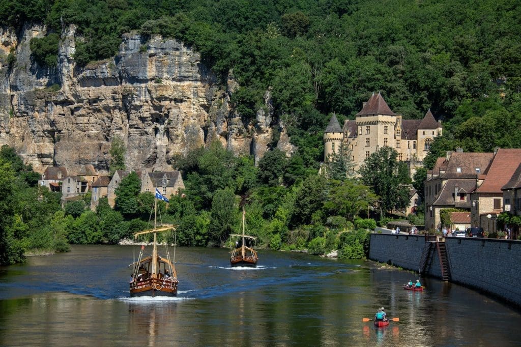A photorealistic image of a medieval castle perched on a cliff overlooking a river in the Dordogne region of France. Two traditional boats sail down the river, while a kayaker paddles alongside them