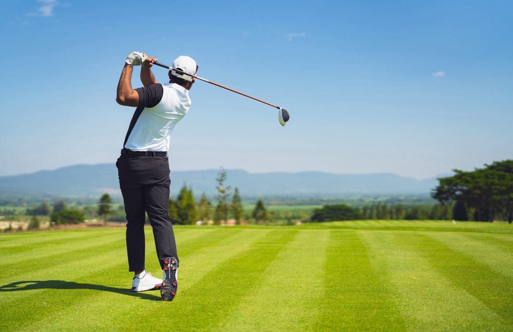 Golfer in a white shirt finishing a swing on a lush, well-maintained golf course with mountains in the distance.