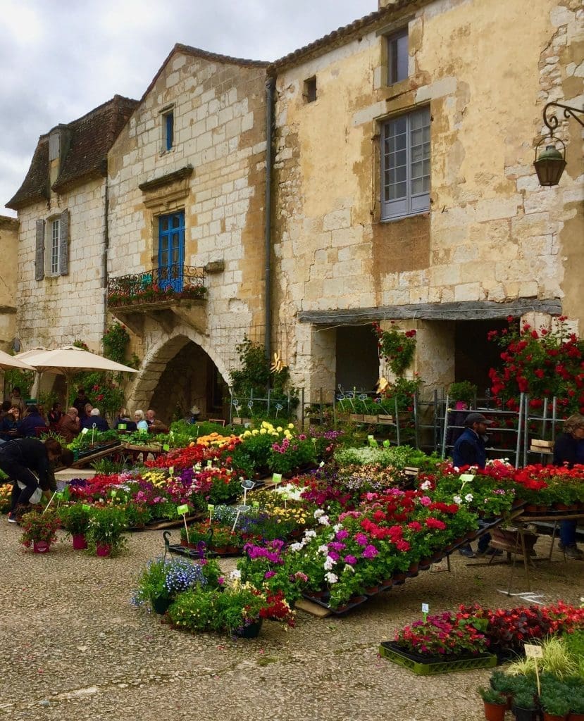 A flower market in Monpazier, France, with colorful flowers and stone buildings in the background.